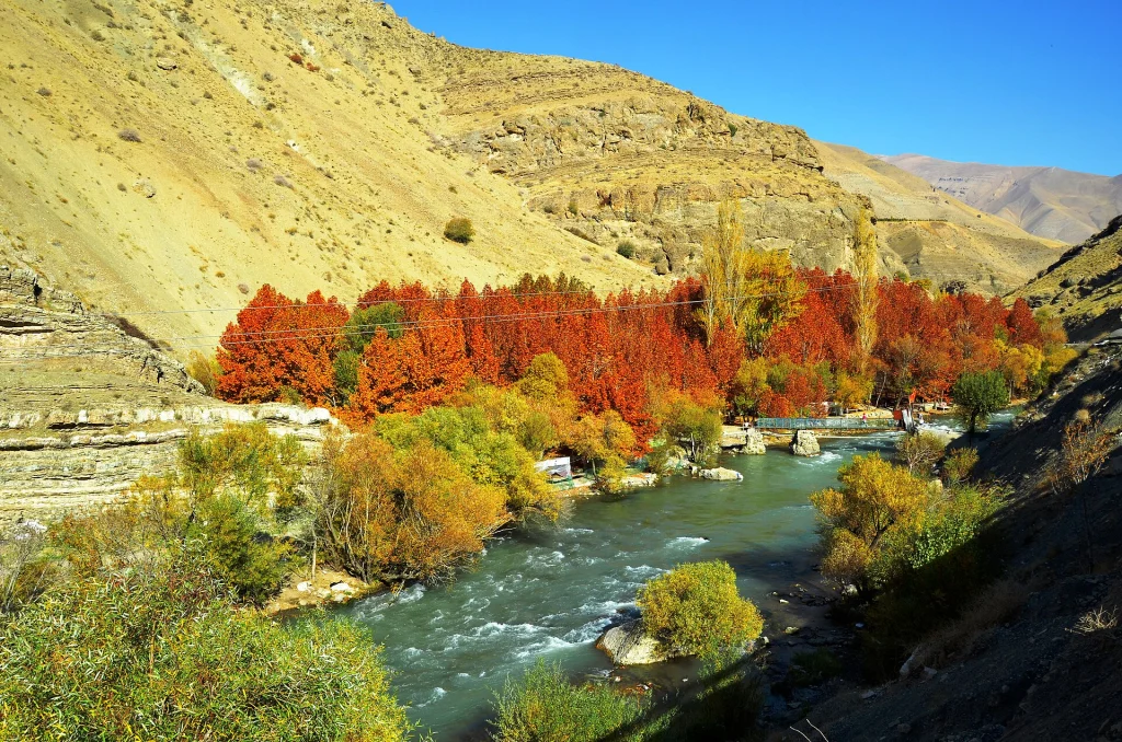 درختان سرخ و نارنجی پاییزی کنار رودخانهٔ کرج در ابتدای جادهٔ چالوس، با یک پل عابر فلزی روی رود و دامنه‌های خشک البرز زیر آسمان آبی
