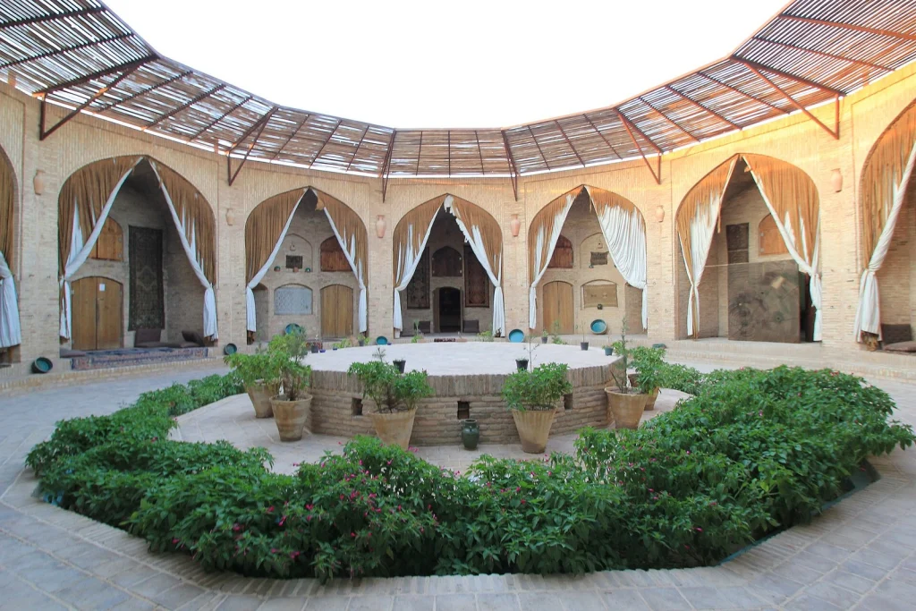 حیاط دوازده‌ضلعی کاروانسرای زین‌الدین نزدیک یزد؛ ایوان‌های طاق‌دار با پرده‌های سفید و درهای چوبی حجره‌ها، سکوی آجری وسط حیاط، گلدان‌ها و باغچهٔ سبز زیر سایه‌بان‌های چوبی