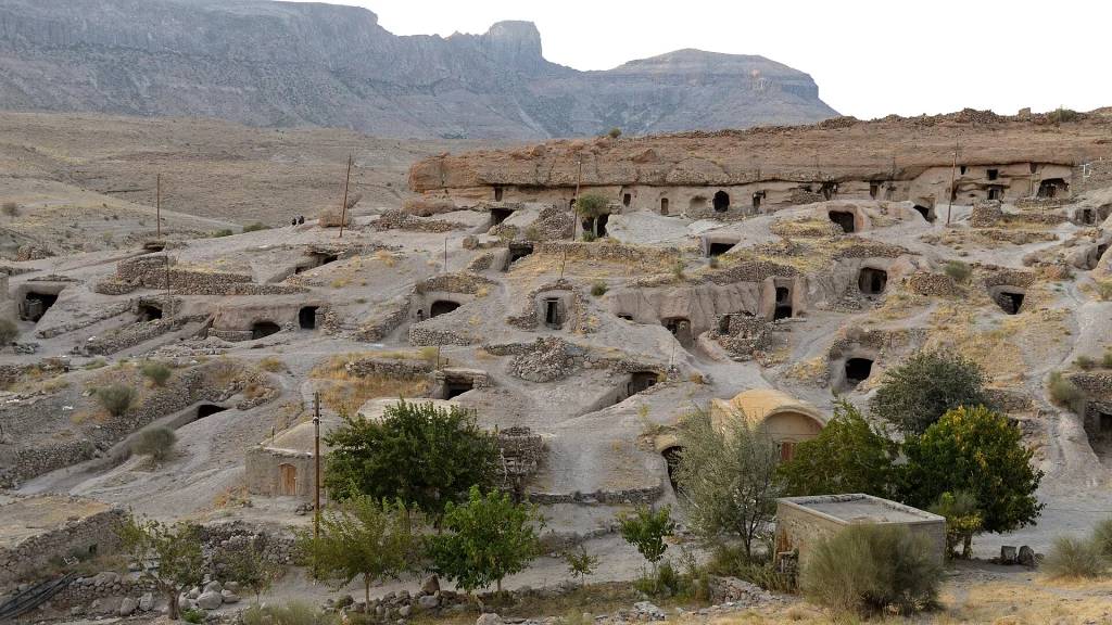 نمای روستای صخره‌ای میمند در شهربابک کرمان: ده‌ها درِ خانهٔ دستکند (کیچه) با دیوارچه‌های سنگ‌چین روی شیب دره، ردیفی از دهانه‌های کنده‌شده زیر لبهٔ صخره و کوه‌های بلند پشت روستا